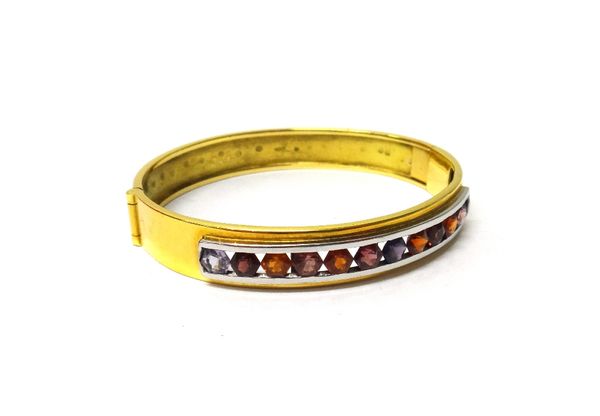 A gold, tourmaline, amethyst and topaz set oval hinged bangle, the front mounted with a row of twelve hexagonal cut, vary coloured gemstones, on a sna