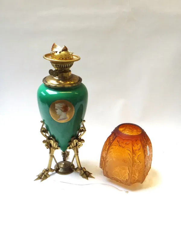 An Aesthetic Movement pottery and brass oil lamp, green ground decorated with portrait roundels, on a triform birds foot cast base, with a later amber