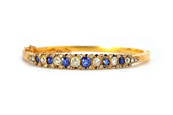 A gold, sapphire and diamond set oval hinged bangle, mounted with a row of alternating cushion shaped sapphires and diamonds to the centre, between pa