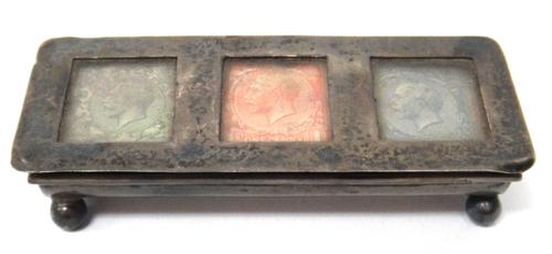 A silver hinge lidded triple compartment postage stamps box, glazed with three differing George V stamps and raised on four feet, the hallmark rubbed.