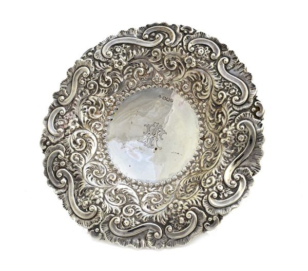 A late Victorian silver shaped circular dish, with floral, foliate and scroll embossed decoration, monogram engraved to the centre, raised on four fee