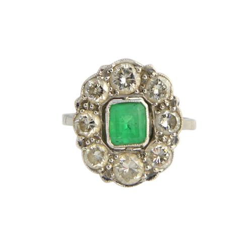 A white gold, emerald and diamond oval cluster ring, mounted with the cut cornered step cut emerald at the centre, within a surround of eight circular