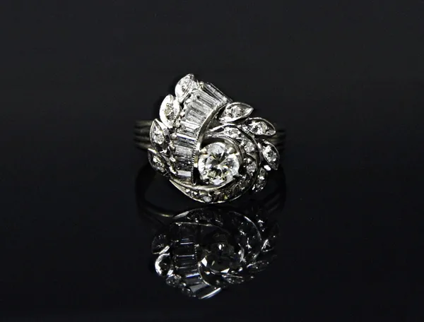 A diamond ring, in a pierced foliate and abstract design, claw set with the principal circular cut diamond at the centre and otherwise mounted with ci