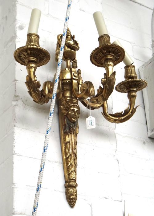A pair of gilt bronze three branch wall appliques, early 20th century, each with urn finial over an acanthus cast backplate issuing three foliate cast