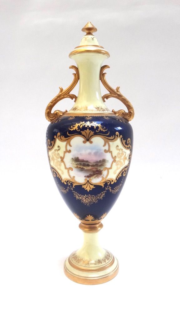 A Coalport porcelain two handled vase and cover, circa 1900, painted with a landscape scene within a gilt scroll cartouche, against a cobalt blue and