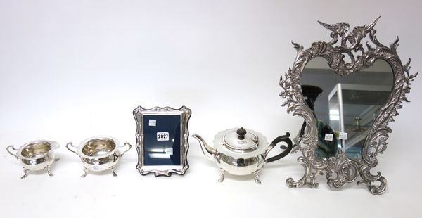 A silver mounted, shaped rectangular photograph frame, a plated three piece tea set and a plated strut backed toilet mirror, pierced in a rococo scrol