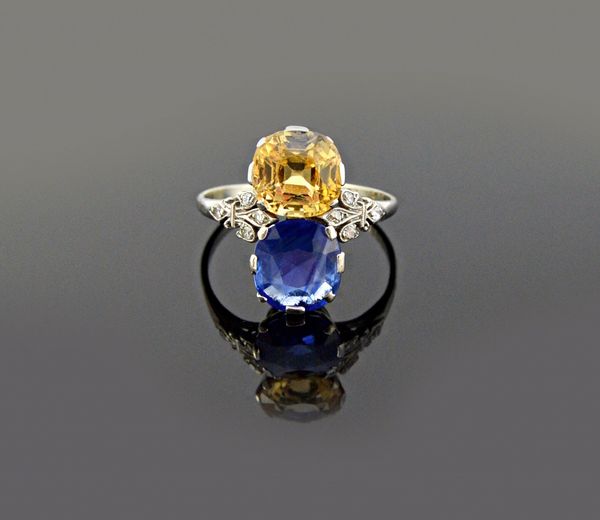 A platinum and white gold ring, claw set with a cushion shaped sapphire and with a cushion shaped yellow sapphire, between diamond set trefoil shaped