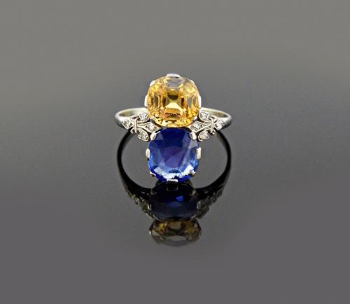 A platinum and white gold ring, claw set with a cushion shaped sapphire and with a cushion shaped yellow sapphire, between diamond set trefoil shaped
