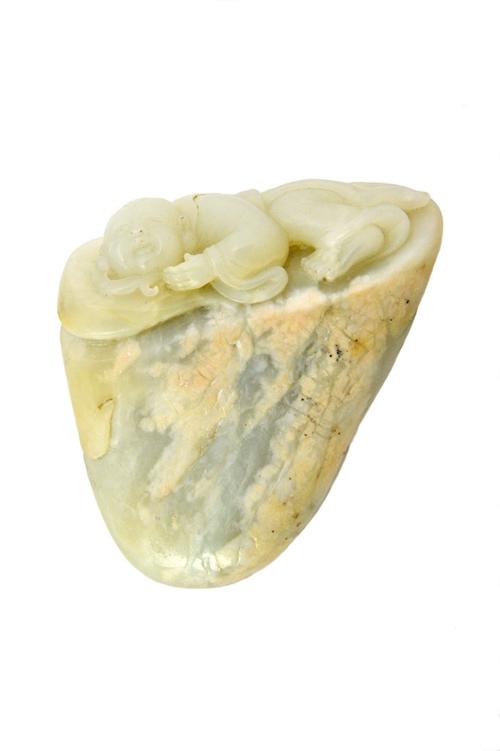 A Chinese jade carving of Budai, depicted lying asleep on a boulder, holding a leaf in his hand, the stone of very pale green tone with mottled white