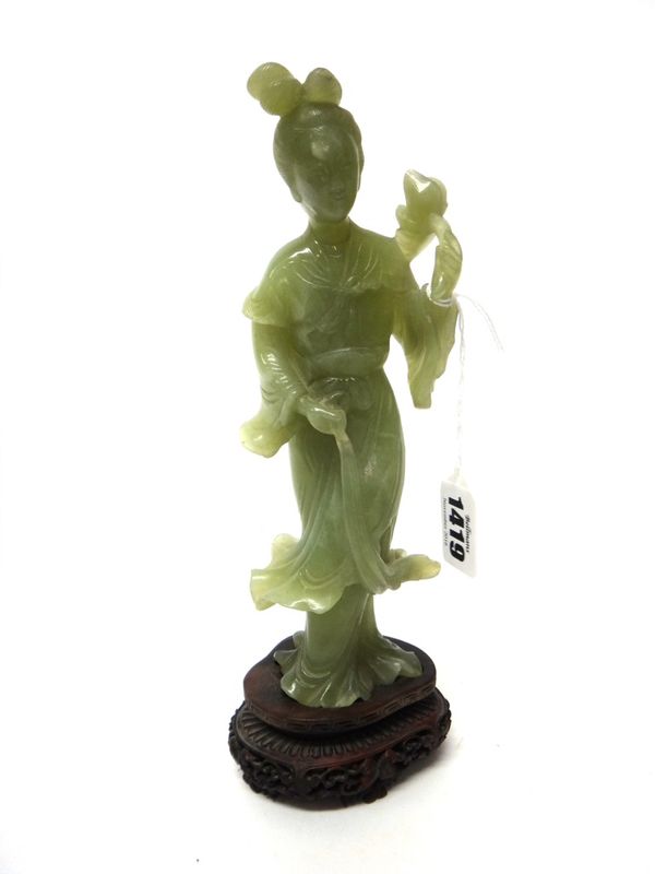 A Chinese celadon jade figure of a woman, late 19th/20th century, standing in long robes holding a lotus flower in one hand, 22cm.high, wood stand.