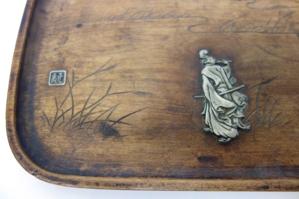 A Japanese wood tray, Meiji period, of rounded rectangular form, inlaid in silver coloured metal with a warrrior walking in a landscape playing pipes,