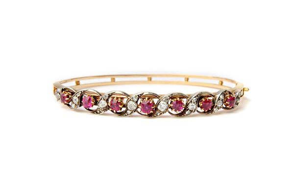 A gold and silver set, ruby and diamond set oval hinged bangle, the front pierced in a scrolling design, mounted with a row of seven cushion shaped ru