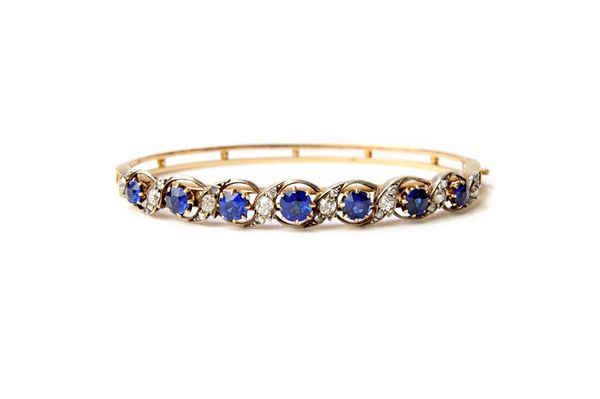 A gold and silver set, sapphire and diamond set oval hinged bangle, the front pierced in a scrolling design, mounted with a row of seven cushion shape