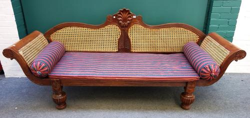 An Anglo-Indian carved hardwood and cane work day bed, the shaped back centred by stylized shell, flanked by out swept arms, on reeded bulbous support