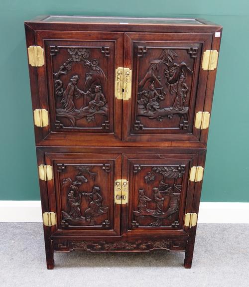 An early 20th century Chinese hardwood double height side cabinet, with two pairs of carved panel doors depicting figures in a landscape, on block sup
