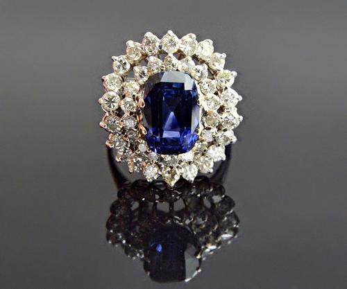 A sapphire and diamond cluster ring, claw set with the cushion shaped sapphire at the centre, in a two tier surround of circular cut diamonds, ring si