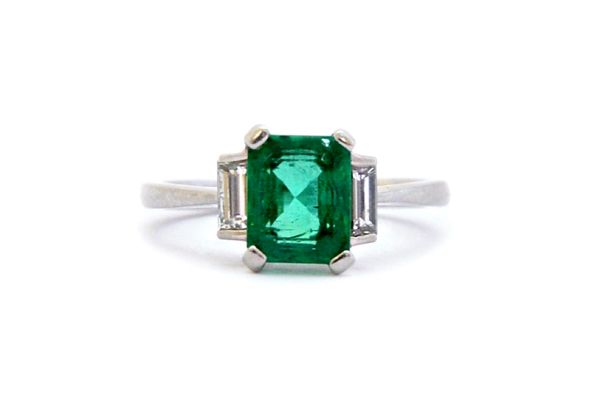 A platinum, emerald and diamond set ring, claw set with the cut cornered rectangular step cut emerald at the centre, between two baguette diamonds, th