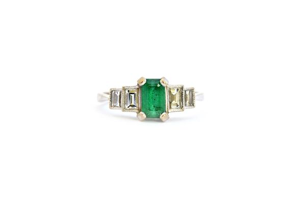 A white gold, emerald and diamond ring, claw set with a rectangular step cut emerald at the centre, between rectangular cut diamond set two stone grad