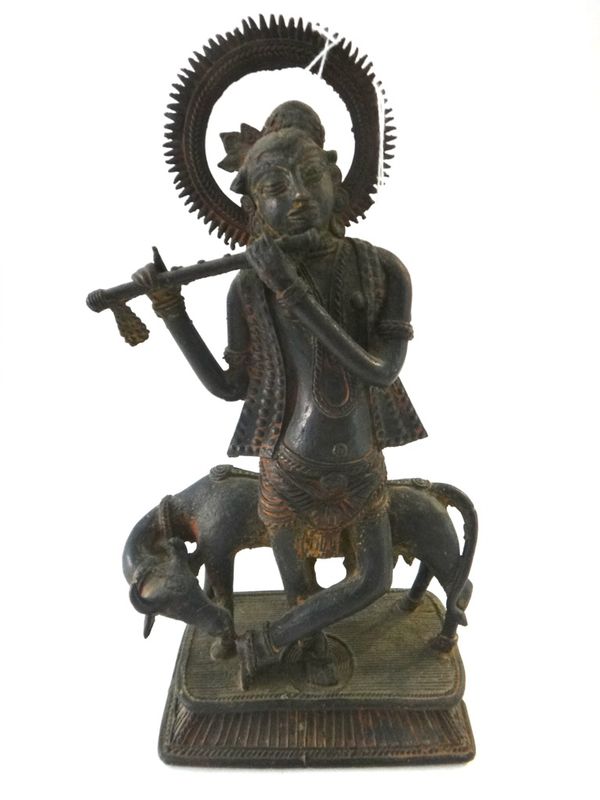 An Indian bronze group of Krishna as a cowherd, the deity standing before a cow and playing the flute, on a rounded rectangular base, 29cm.high.