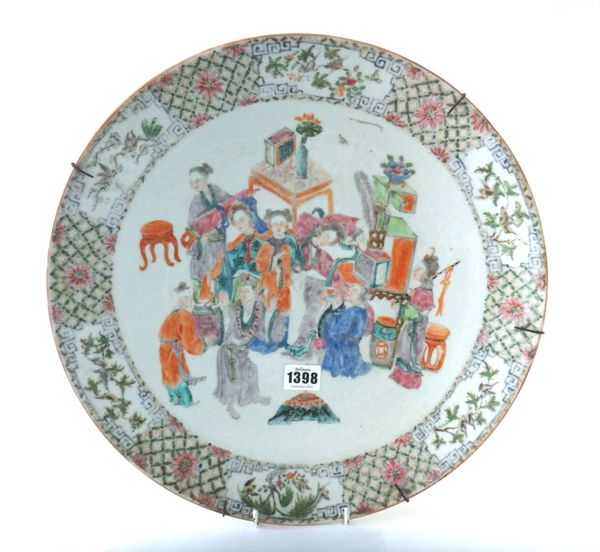 A Chinese famille-rose circular dish, circa 1900, painted with figures at leisure amongst tables, chairs and vases, beneath a diaper pattern border re