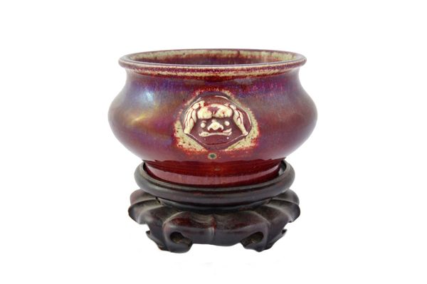 A Chinese flambé glazed censer, 18th/19th, of compressed form set with lion mask handles, 13.5cm. wide across handles, wood stand. Illustrated