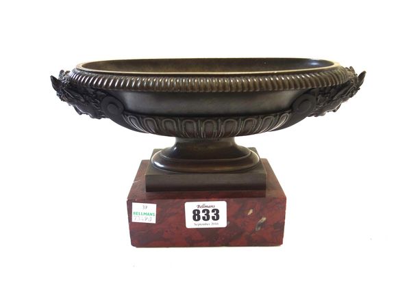 After the Antique; a bronze oval urn of shallow form, 19th century, the twin handles cast as Bacchus masks, on a rouge polished marble plinth, 23.5cm