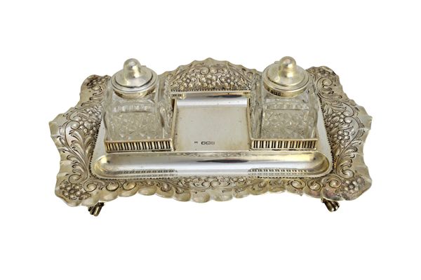 A late Victorian silver twin bottle inkstand, of shaped rectangular form, the borders with floral and scroll embossed decoration, raised on four pierc