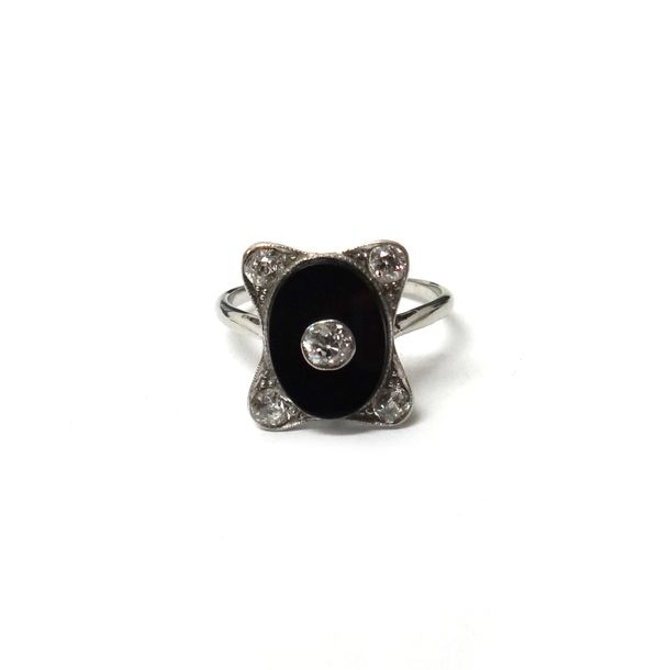 A platinum, diamond and black onyx ring, in a curved rectangular panel shaped Art Deco design, centred by a collet set cushion shaped diamond, on an o