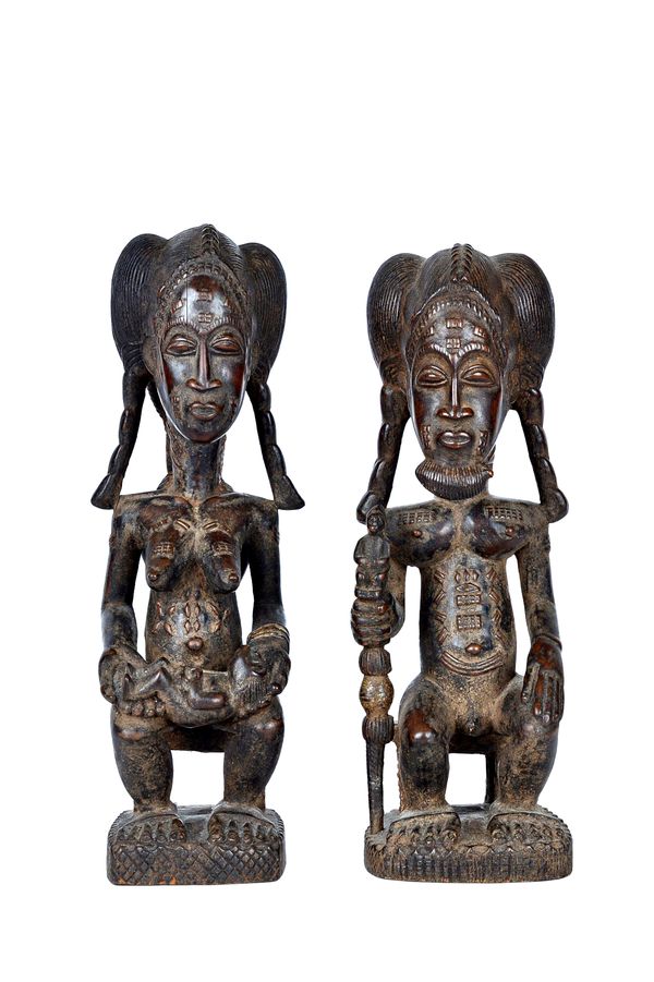 A pair of African carved wooden tribal figures, mid-20th century, both seated, the man holding a staff, the lady, breasts bared holding a small child,