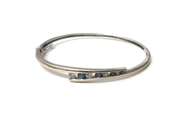 A white gold, diamond and treated blue diamond set oval hinged bangle, the front mounted with a row of alternating circular cut diamonds and circular