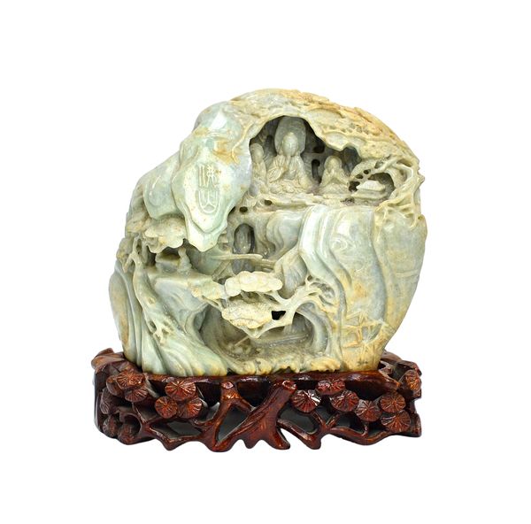 A Chinese jade boulder carving, of pale celadon tone with light brown and cream  inclusions, carved with buddha seated in a grotto flanked by two acol