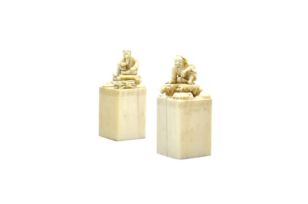 A pair of Chinese ivory seals, late 19th century, of square section with key pattern borders, each surmounted by a figure seated on a rock, one with a