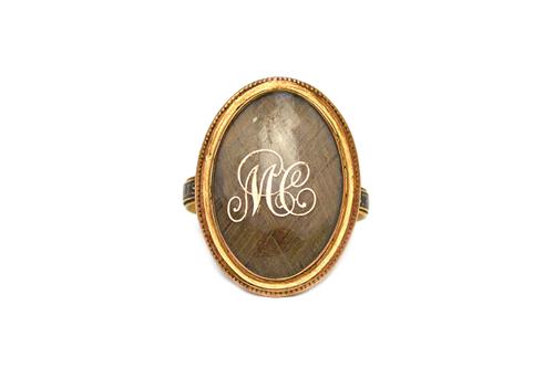A George III gold and black enamelled mourning ring, the oval bezel glazed with a woven hair locket compartment and with the gilt script initials 'M C