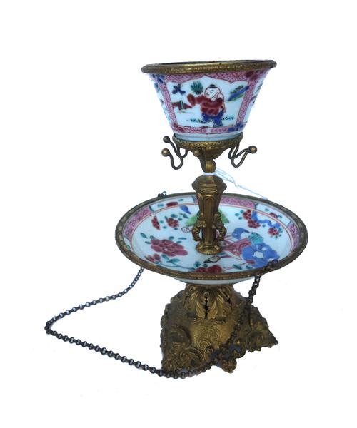 A Chinese tea bowl and saucer, early Qianlong,  with gilt metal mounts, arranged over two tiers linked with chains on a foliate cast triform base, 17c