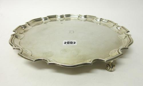 A silver shaped circular salver having a piecrust rim, in the Chippendale style, engraved with the initial D at the centre, raised on three scrolling