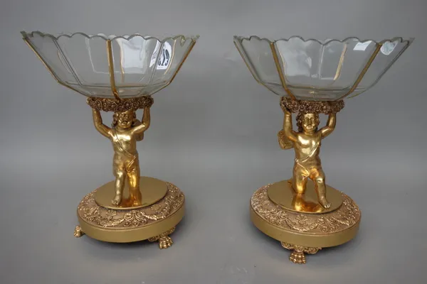 A pair of gilt metal and glass mounted figural tazza, each putto figure supporting a cut glass bowl of segmented form, over a foliate cast circular ba