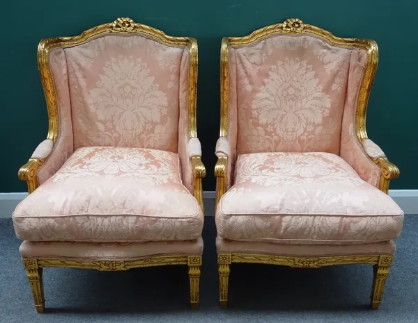 A pair of gilt framed wingback armchairs in the Louis XV/XVI transitional period style, each with serpentine seat on tapering supports, 71cm wide x 10