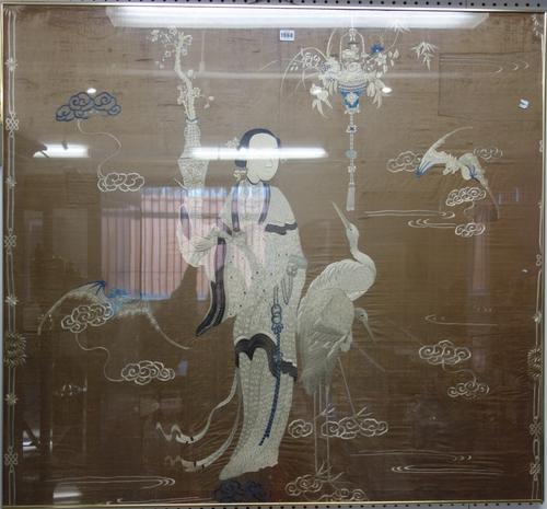 A Chinese silk panel, late 19th/early 20th century, worked with a woman standing holding a vase with two cranes beside her, 109cm. by 119 cm., (a.f),