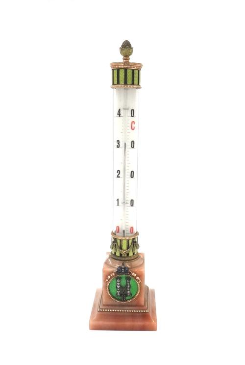 A gold and enamel mounted pink hardstone table thermometer of column form, raised on a square base, the base applied with two diamond set and enamelle