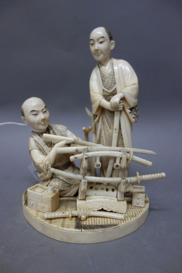 A Japanese sectional ivory okimono of swordmakers, Meiji period, one seated on a mat inspecting a long sword, another standing beside, a stand and swo