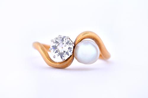 A gold, diamond and cultured pearl set ring, claw set with a cushion shaped diamond and with a cultured pearl, in a crossover design, ring size L and