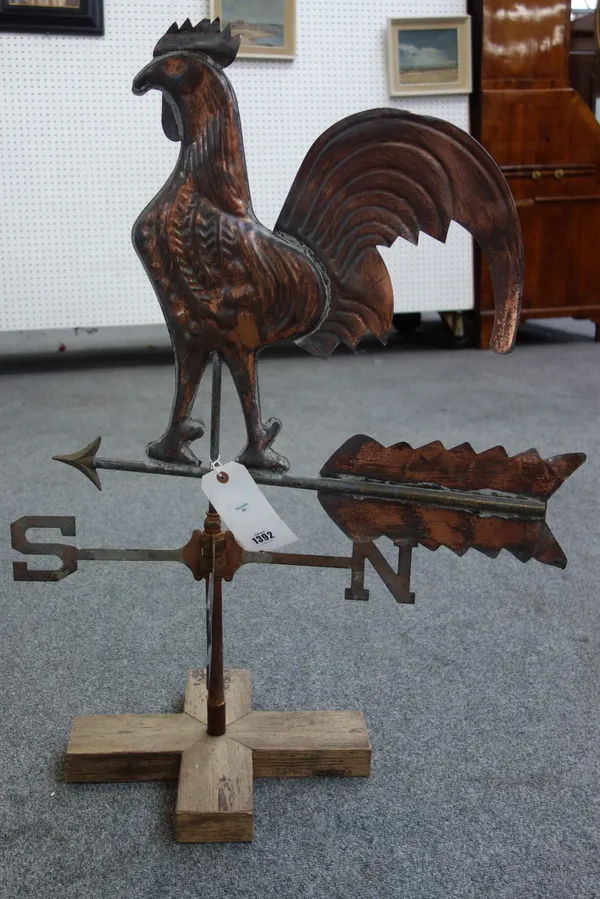 A copper coloured metal weather vane with Cockerel finial over an arrow and directional pointers, mounted on a wooden base. 83cm high.