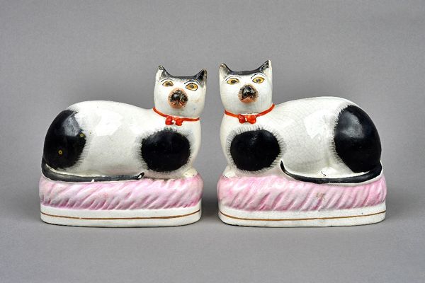 A pair of Staffordshire pottery cats, second half 19th century, each painted with black patches and wearing orange collars, modelled recumbent on an o