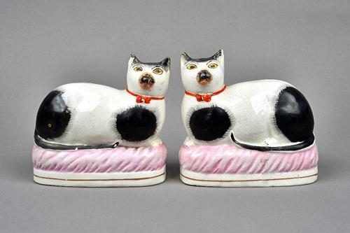 A pair of Staffordshire pottery cats, second half 19th century, each painted with black patches and wearing orange collars, modelled recumbent on an o