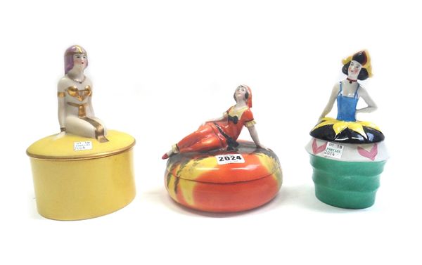 Three Continental porcelain boxes and covers, each of Art Deco polychrome form with a female figure to the lid, the tallest 18.5cm high (a.f), (3).