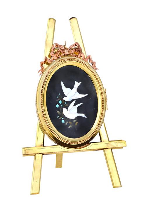 A brass pietra dura folding photograph frame, circa 1900, depicting two doves in flight, in an oval hinged frame, mounted on a strut back easel frame,