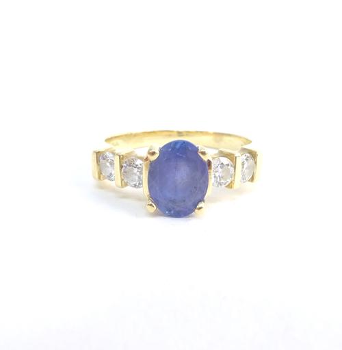 A gold, diamond and pale blue / lilac coloured gem set ring, claw set with the oval cut blue / lilac coloured gem at the centre, between circular cut