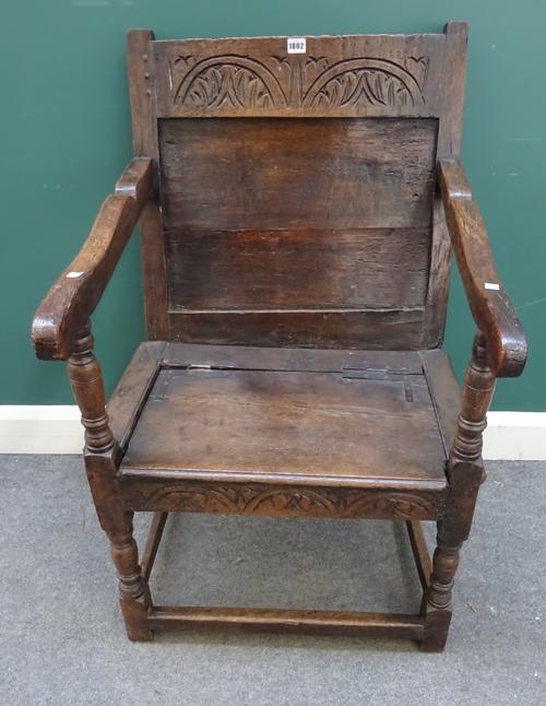An 18th century and later carved oak country armchair, the low back with lunette carved top, hinged seat within nocked arms, turned legs united by str