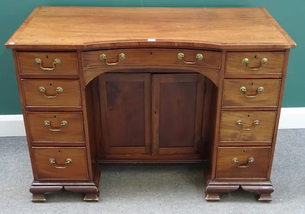 A George III crossbanded mahogany kneehole writing desk, the convex top above nine drawers about the central cupboard, on ogee bracket feet, 121cm wid