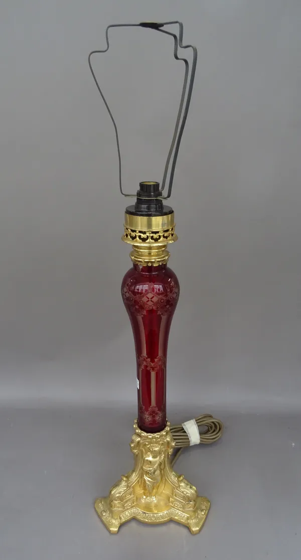 A Victorian style gilt metal and etched ruby glass table lamp, the triform base cast with a putto playing a mandolin, with shade, 50cm high excluding